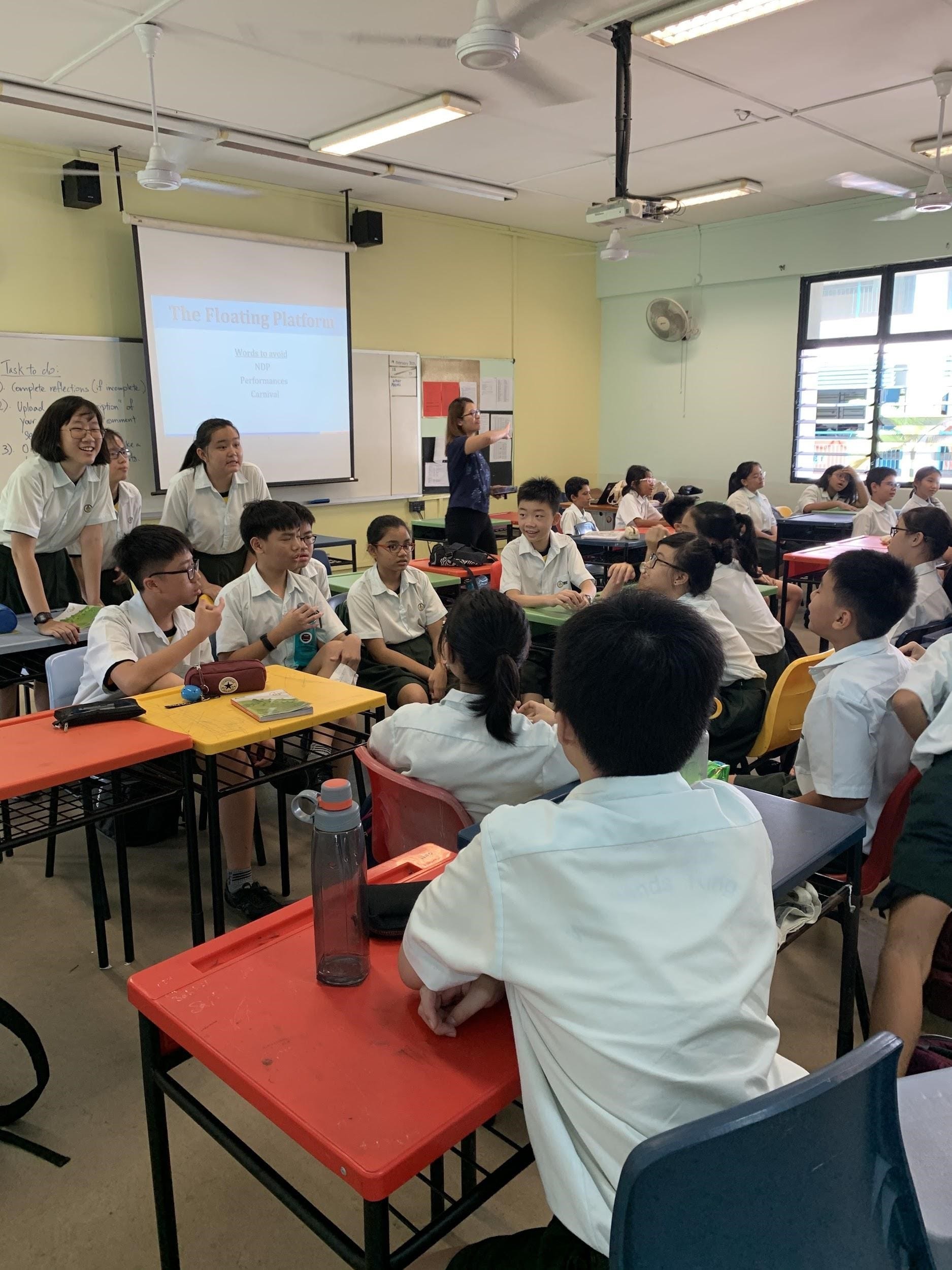 Students in white uniforms sit at colorful desks in a classroom while a teacher points at a projected presentation screen.